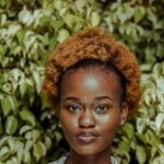 Portrait of a young woman with an afro hairstyle in front of vibrant green foliage.